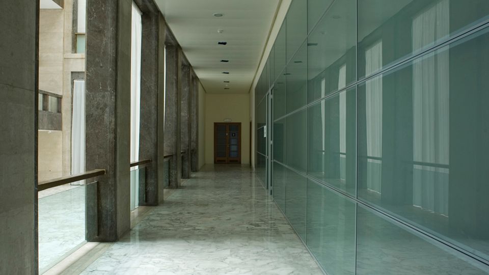 Corridoio interno di un edificio moderno con pavimento in marmo chiaro, colonne in pietra e grandi finestre sul lato sinistro. A destra una lunga parete vetrata con pannelli e telai metallici, con porta in fondo al corridoio.