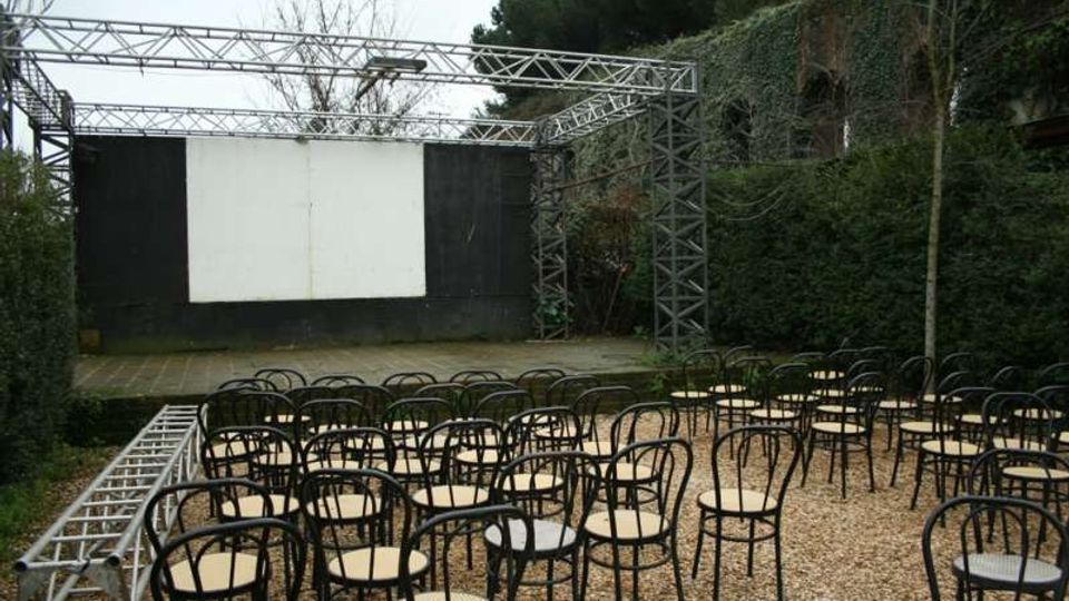 Piccolo teatro all’aperto con palco e grande schermo bianco incorniciato da pannelli neri e struttura metallica a traliccio. Davanti al palco ci sono file di sedie in metallo su ghiaia, circondate da siepi e vegetazione.