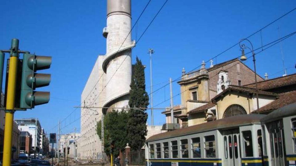 Strada urbana con binari e linee elettriche del tram, con un tram blu in primo piano. Sullo sfondo si vede un alto edificio industriale con grande ciminiera cilindrica e rampa elicoidale esterna, accanto a edifici storici con tetti in coppi e facciate in muratura.