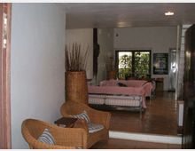 Interno di un’area lounge con pavimento in cotto e pareti chiare, arredata con poltrone in vimini, tavolini e grandi vasi decorativi. Sul fondo si vede un divano coperto in tessuto rosa e una porta a vetri che dà su un’area verde esterna.
