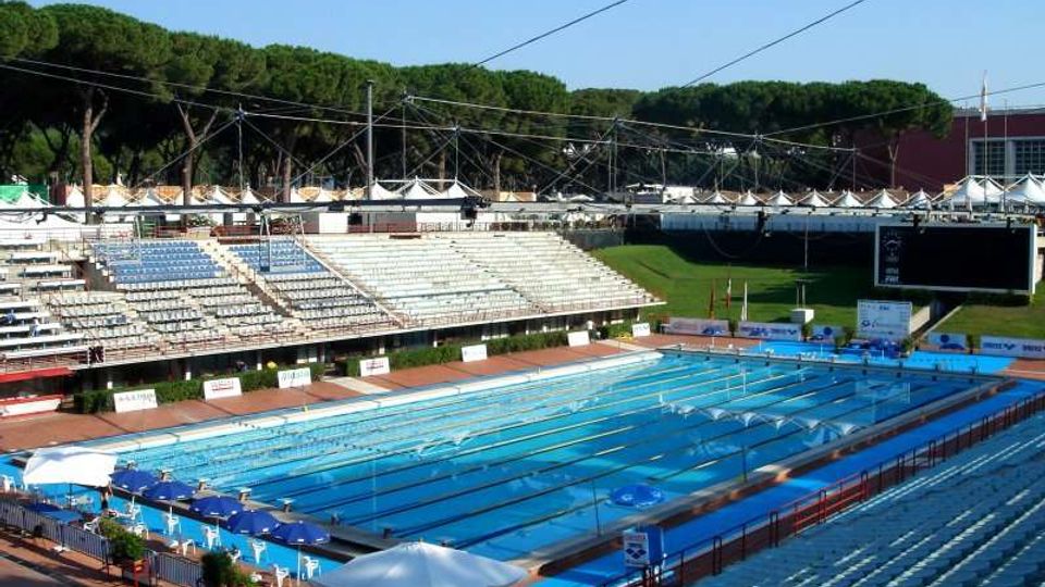 Piscina olimpionica all’aperto con corsie delimitate, circondata da grandi tribune in cemento e sedute. Sullo sfondo si vedono cavi e strutture di copertura, un grande schermo e una fascia di alberi.
