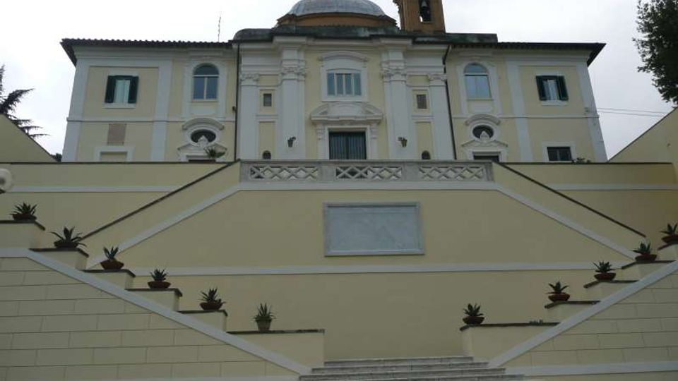 Facciata di un edificio religioso in stile classico con cupola e campanile, vista dal basso. In primo piano una lunga scalinata in pietra con balaustre e terrazze laterali decorate da vasi di piante.