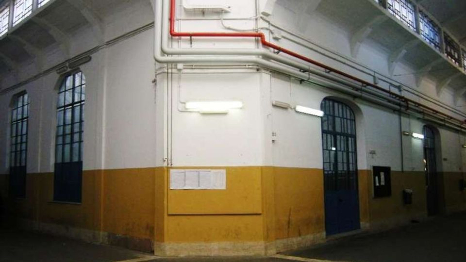 Interno di un capannone industriale con soffitto alto e travi a volta, grandi finestre ad arco e porte metalliche blu lungo le pareti. Pareti bianche e ocra con tubazioni a vista rosse e grigie vicino al soffitto e pavimento in cemento segnato.