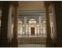 Cortile interno di un edificio storico con colonne in pietra e ballatoio con balaustra, affacciato su una facciata con arcate e finestre ad arco. Al centro si vede una porta in legno e pareti dai toni chiari sotto un soffitto alto.