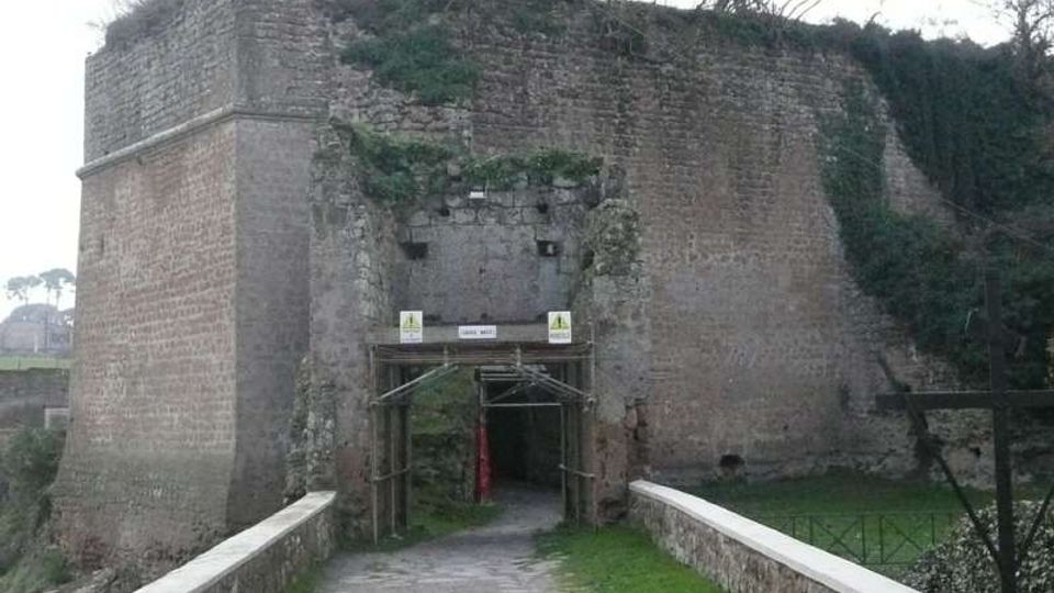 Ingresso ad arco in una massiccia fortificazione in muratura di mattoni e pietra, parzialmente coperta da vegetazione, raggiunto da un vialetto su un piccolo ponte con muretti laterali. Intorno si vede un’area verde con alberi spogli e pendii erbosi.
