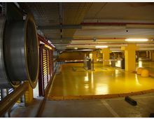Parcheggio sotterraneo con pavimento giallo lucido e colonne gialle, illuminato da luci al neon e con tubazioni e canaline a soffitto. Si vedono una sbarra di accesso, segnaletica con frecce sulla pavimentazione e una grande ventola industriale sulla sinistra.