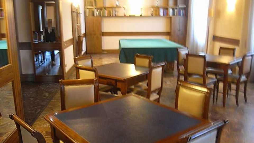 Sala interna tipo lounge o sala da tè con tavoli e sedie in legno, pavimento in marmo e soffitto con pannelli in legno. Sul fondo ci sono scaffali con libri e un tavolo da biliardo coperto, con una grande finestra laterale che illumina l’ambiente.