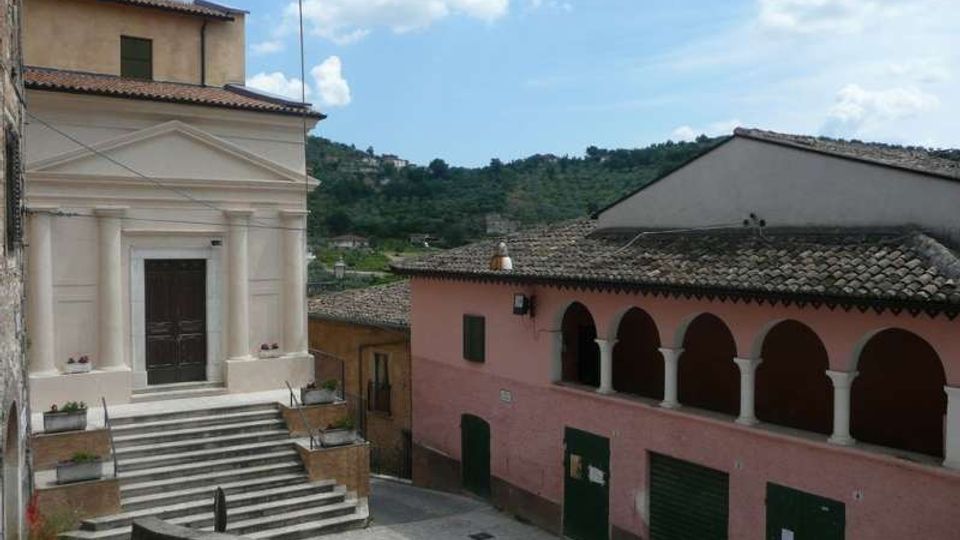 Piazza in un borgo collinare con una chiesa chiara a facciata classica e scalinata in pietra. Di fronte si vede un edificio rosa con portico ad archi e tetto in coppi, sullo sfondo colline verdi e cielo parzialmente nuvoloso.
