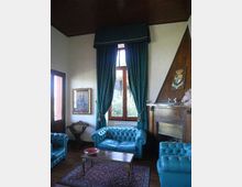 Salotto con divani Chesterfield in pelle azzurra, tavolino in legno su tappeto decorato e grande finestra con tende pesanti verde scuro. A destra c’è un camino rivestito in legno con cappa triangolare e stemma, con soffitto in legno e pareti chiare.