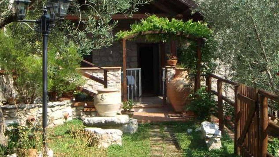 Ingresso di una casa rustica in pietra con tetto in legno, circondata da ulivi e vegetazione. Un sentiero erboso conduce a un pergolato coperto di rampicanti, con vasi decorativi, lampioni e una staccionata in legno lungo il lato destro.