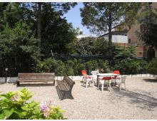 Cortile-giardino con area in ghiaia, due panchine in legno e un tavolo con sedie in plastica bianche e rosse. Sullo sfondo ci sono alberi e arbusti fitti, con un edificio chiaro parzialmente visibile dietro la vegetazione e una rete di recinzione.