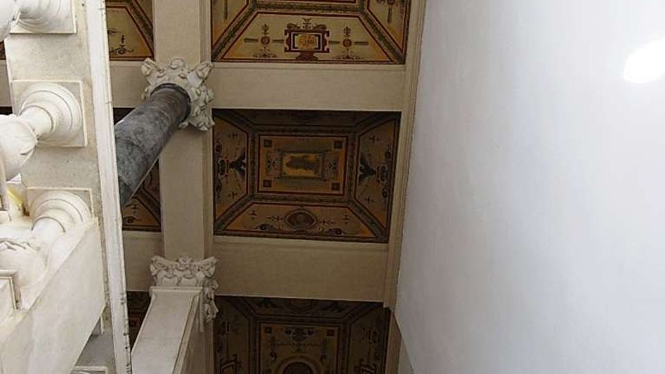 Soffitto interno decorato con riquadri a cassettoni dipinti con motivi geometrici e medaglioni centrali, sostenuti da travi e colonne. A sinistra si vede una balaustra in stile classico, mentre a destra una parete bianca corre lungo un vano stretto verso l’alto.