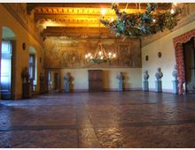 Ampia sala storica con pavimento in pietra e soffitto a travi lignee decorato, illuminata da grandi lampadari sospesi. Lungo le pareti sono disposti busti su piedistalli e un grande affresco, con finestre e porte laterali incorniciate da tendaggi.