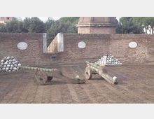 Cortile in mattoni con due carri/cataste in legno che trasportano pile di palle di cannone in metallo. Sullo sfondo si vede un alto muro in laterizio con targhe ovali e una struttura circolare in muratura, circondata da alberi.