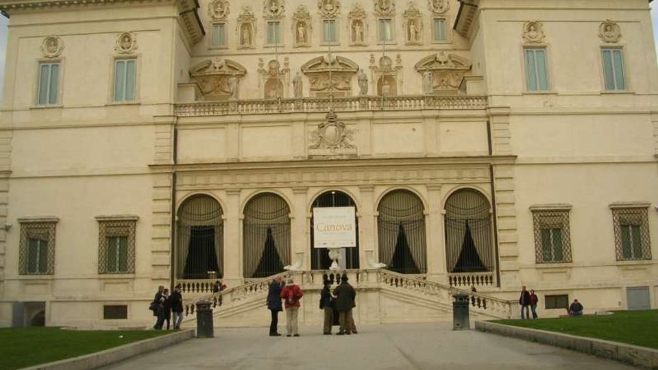 Facciata di un grande palazzo storico in stile rinascimentale, con decorazioni scolpite, finestre simmetriche e tre ampie arcate al piano terra. Una scalinata centrale conduce all’ingresso principale, affacciato su un viale pedonale con prati ai lati.