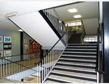 Atrio interno di un edificio con ampia scala in cemento e ringhiere metalliche bianche e nere, illuminato da plafoniere a soffitto. Sulle pareti del piano inferiore si vedono quadri e un grande murale colorato, con finestre sul fondo.