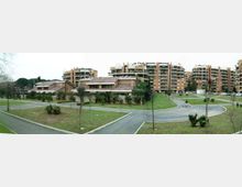 Panorama di un complesso residenziale con alti condomini in mattoni e ampi balconi, affiancati da edifici più bassi con tetti a falde. In primo piano si vedono strade interne asfaltate e aree verdi con prati e alberi giovani, disposte tra i percorsi.
