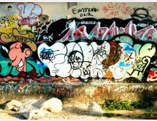 Muro di cemento all’aperto ricoperto da graffiti e scritte colorate, con grandi forme a lettere e un disegno in stile fumetto. Alla base del muro si vedono un marciapiede in cemento, ghiaia e alcune piante basse.