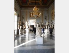 Sala museale classica con pareti ornate e cornicioni decorativi, illuminata da un grande lampadario in vetro e con pavimento in marmo a motivi geometrici. Busti e statue in marmo sono disposti su mensole lungo le pareti e una statua su piedistallo occupa il centro della stanza.