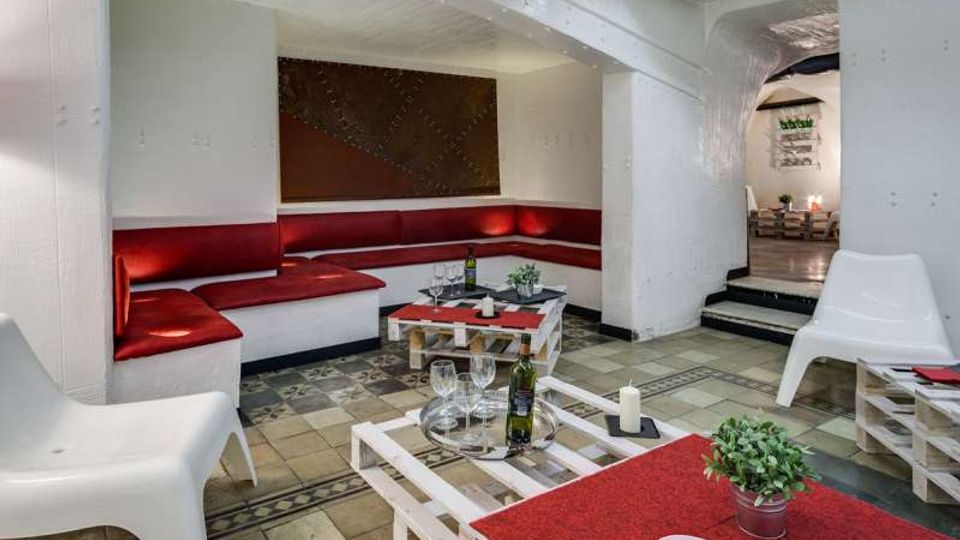 Salotto interno in stile lounge con pareti e soffitto bianchi, divano ad angolo con cuscini rossi lungo le pareti e pavimento a piastrelle decorative. Tavolini bassi in legno tipo pallet con bottiglie e bicchieri al centro e, sullo sfondo, un passaggio ad arco che conduce a un’altra sala.