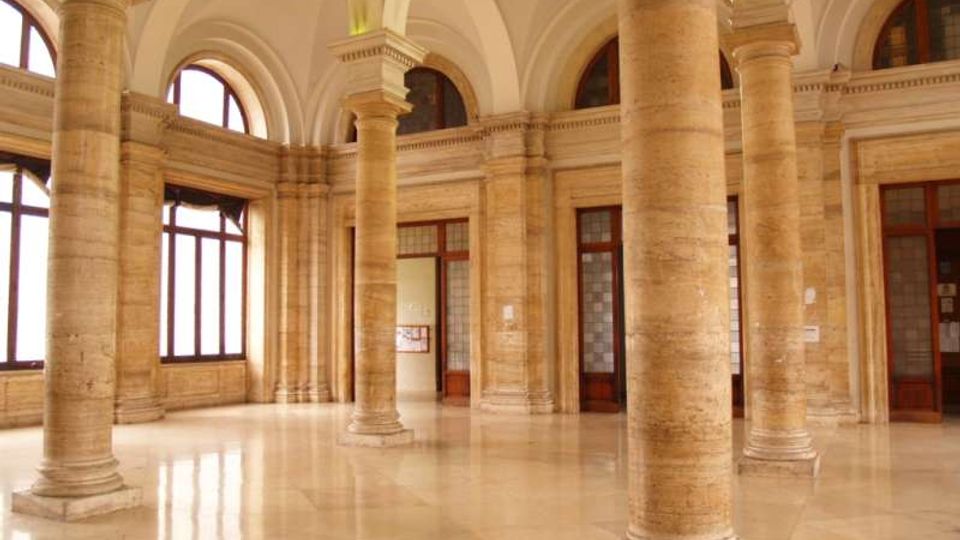 Interno di un ampio atrio in stile classico con colonne in pietra, pavimento lucido e soffitto a volte. Grandi finestre ad arco e porte vetrate lungo le pareti illuminano lo spazio.