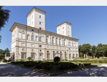 Grande palazzo storico in stile rinascimentale con facciata chiara decorata da cornici e finestre regolari, affiancato da due torri rettangolari. In primo piano si vede un giardino geometrico con siepi basse e vialetti, circondato da alberi sotto un cielo sereno.