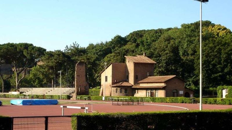 Impianto sportivo all’aperto con pista di atletica rossa e area per il salto in alto, recintato e con un alto palo per l’illuminazione. Sullo sfondo si vedono edifici in mattoni con tetti in coppi e una torre, circondati da alberi e vegetazione.