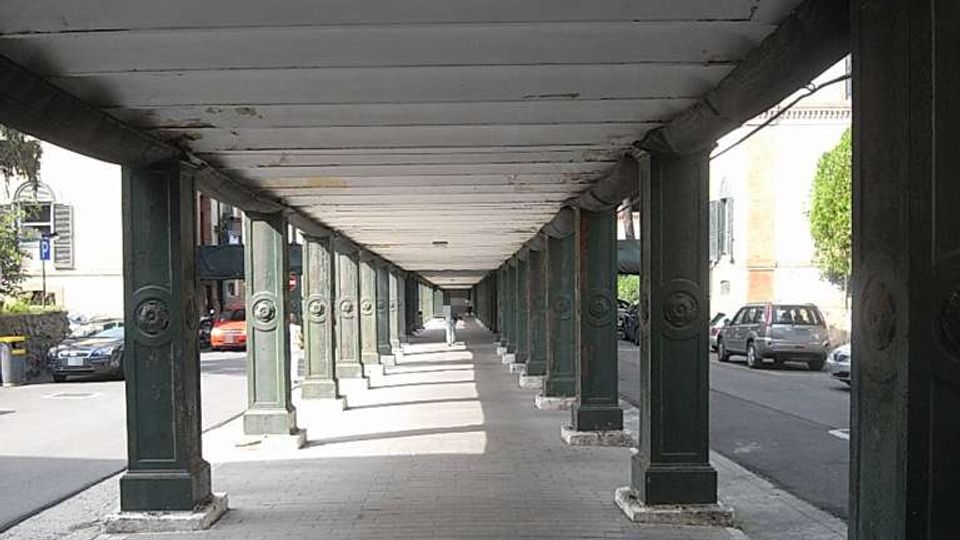 Portico urbano lungo e rettilineo con pavimentazione in pietra e soffitto a travi, sostenuto da una fila di colonne in metallo scuro. Ai lati si intravedono la strada con auto parcheggiate e alcuni edifici.
