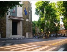 Facciata di un edificio pubblico in pietra con grande portale d’ingresso e bandiere su pennoni, affacciato su una strada con strisce pedonali. Accanto si vede un incrocio alberato con alti alberi e palazzi sullo sfondo.
