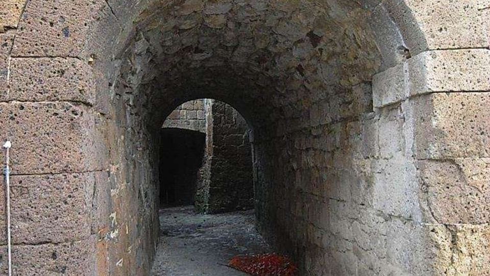 Passaggio stretto in pietra con grande arco in muratura e soffitto voltato, che conduce a un corridoio ombreggiato con pavimento in ciottoli. Le pareti sono in blocchi di pietra irregolari, con un secondo arco visibile in fondo.