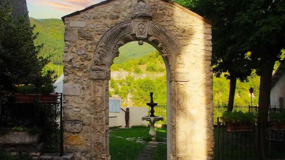 Antico portale in pietra con arco scolpito e decorazioni, che introduce a un piccolo giardino. Oltre l’arco si vede una fontana centrale e, sullo sfondo, colline verdi con alberi e un cielo al tramonto.