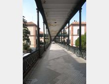 Passerella pedonale sopraelevata con lunga copertura e colonne in metallo, pavimentazione a piastrelle e ringhiere laterali. Ai lati si vedono edifici in muratura con finestre alte e tetti in tegole, con alberi e auto nel cortile sottostante.