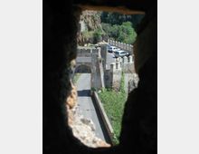 Veduta dall’interno di una feritoia su mura fortificate in pietra con merli e un arco d’ingresso che attraversa una strada. Sullo sfondo si vede una curva della carreggiata lungo un costone roccioso con vegetazione e alberi.