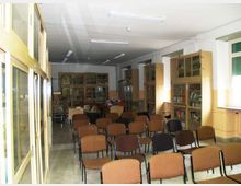 Aula interna con file di sedie imbottite disposte a platea, illuminata da lampade fluorescenti a soffitto. Lungo le pareti ci sono alte librerie e armadi vetrati con libri e materiali, con finestre sul lato destro.