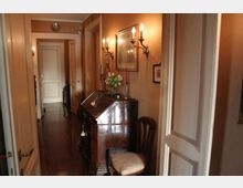 Corridoio interno stretto con pareti color beige, pavimento in legno scuro e porte bianche lungo il passaggio. Sul lato destro ci sono applique a parete, quadri e un mobile scrittoio in legno con fiori e candelabri, affiancato da una sedia.