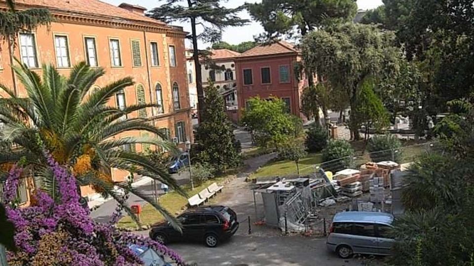 Veduta dall’alto di un cortile con parcheggio e aiuole, circondato da edifici in mattoni dai tetti in tegole. Sono presenti alberi alti, una palma e fioriture colorate, oltre ad alcune aree con materiali e strutture da cantiere.