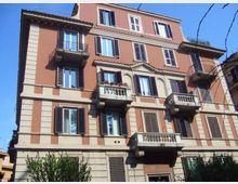 Facciata di un palazzo residenziale di cinque piani con intonaco color terracotta e cornici chiare, finestre con persiane scure e più balconi sporgenti con ringhiere in ferro e balaustre. L’edificio è ripreso dal basso contro un cielo azzurro, con alcuni rami e vegetazione visibili ai margini dell’inquadratura.