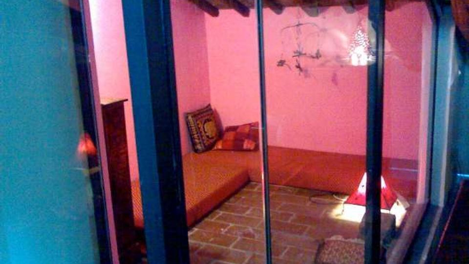 Interno di una piccola stanza con pareti rosa e soffitto con travi in legno a vista, pavimento in mattonelle e un letto basso con cuscini. La scena è ripresa attraverso una vetrata con montanti verticali scuri.