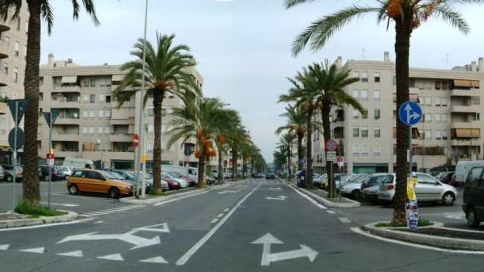 Viale urbano ampio con corsie e frecce direzionali sull’asfalto, fiancheggiato da palme e auto parcheggiate ai lati. Sullo sfondo si vedono palazzi residenziali moderni con balconi lungo entrambi i lati della strada.