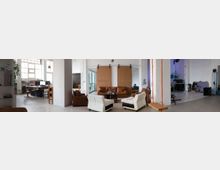 Ampio loft adibito a studio con grandi finestre industriali, pareti bianche e colonne, che include una zona ufficio con scrivanie e computer e un’area lounge con divani e poltrone. Sullo sfondo si vedono attrezzature e arredi da studio in un ambiente aperto e luminoso.