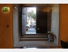 Ingresso interno con porta vetrata e gradini in pietra che conducono all’uscita, affiancata da una parete in legno. All’esterno si vedono la strada con auto parcheggiate e alberi; all’interno è presente un cartello di uscita di emergenza e un portaombrelli.