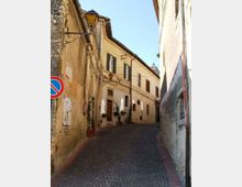 Strada stretta in salita con pavimentazione in ciottoli tra edifici storici in intonaco chiaro, con persiane, porte e vasi alle finestre. Sulla parete a sinistra sono visibili una lanterna a muro e un cartello di divieto di sosta, con cavi sospesi lungo la via.