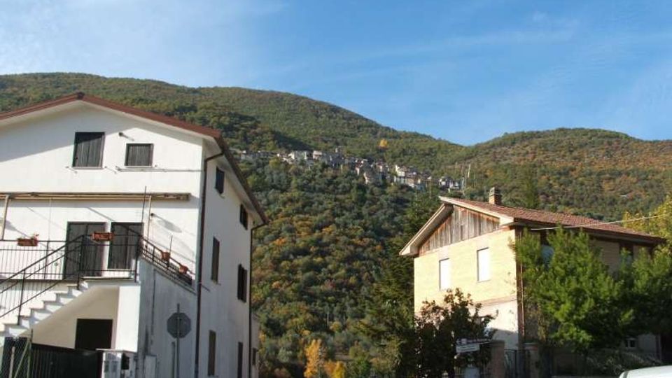 Strada di paese ai piedi di colline boscose, con due case in primo piano (una bianca a più piani con scala esterna e una con tetto a falde) e un borgo arroccato visibile sul crinale in lontananza sotto un cielo sereno.