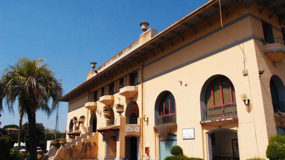 Edificio a due piani color crema con grandi finestre ad arco e balconcini con ringhiere, affacciato su un piccolo piazzale con siepi curate. Sul lato sinistro si vedono una scalinata esterna con vasi ornamentali e una palma, sotto un cielo sereno.