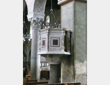 Interno di una chiesa storica con pulpito in marmo decorato da pannelli geometrici e colonne tortili, addossato a un pilastro. Si vedono archi affrescati, colonne in pietra e lampadari sospesi lungo la navata.
