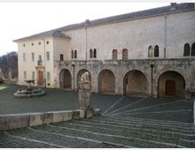 Cortile in pietra di un complesso storico con lunga facciata in muratura chiara e portico ad archi, affacciato su una piazza lastricata con scalinata. In primo piano si vede una piccola fontana circolare e un pilastro in pietra, con edifici a tetto in coppi sullo sfondo.