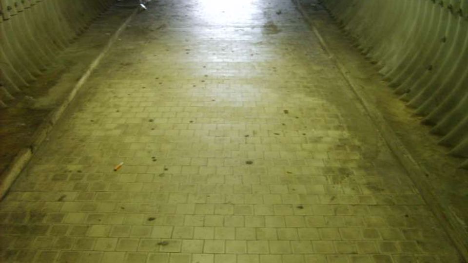Interno di un sottopassaggio pedonale in cemento con pareti laterali ondulate e pavimento in mattonelle, illuminato da luce intensa all’uscita in fondo al tunnel. L’ambiente appare lungo e stretto, con segni di sporco e umidità sul pavimento.
