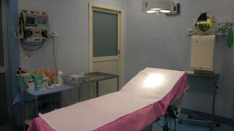 Sala medica/ambulatorio con lettino da visita coperto da lenzuolo bianco e telo rosa, illuminato da una lampada operatoria a soffitto. Sulle pareti sono presenti monitor e apparecchiature mediche, con carrelli e un tavolo con strumenti e materiali.