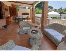Ampio portico coperto di una villa con pavimento in cotto, colonne in mattoni e balaustra bianca, arredato con salotto in vimini e tavolo da pranzo in ferro. Sullo sfondo si vedono il giardino e alcune case, con un grande ombrellone nel cortile esterno.