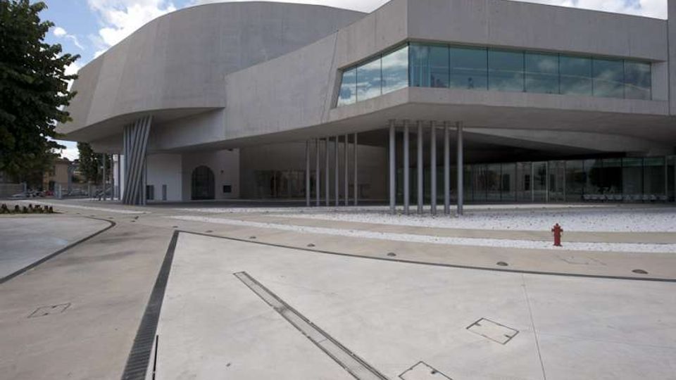 Edificio moderno in cemento grigio con grandi volumi sporgenti e facciata vetrata continua al primo piano, sostenuta da una fila di pilastri sottili. Davanti si apre un’ampia piazza pavimentata in cemento con canalette di drenaggio e pochi elementi urbani, sotto un cielo parzialmente nuvoloso.